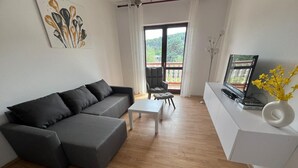 Una sala moderna con un sofá seccional gris, una mesa de centro blanca y un televisor sobre un mueble blanco. Hay un balcón con vistas a zonas verdes.