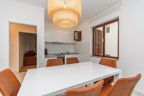 A modern kitchen with white cabinets, a dining table with six chairs, and a large window with wooden shutters. - Apartments Milan (Lopar)