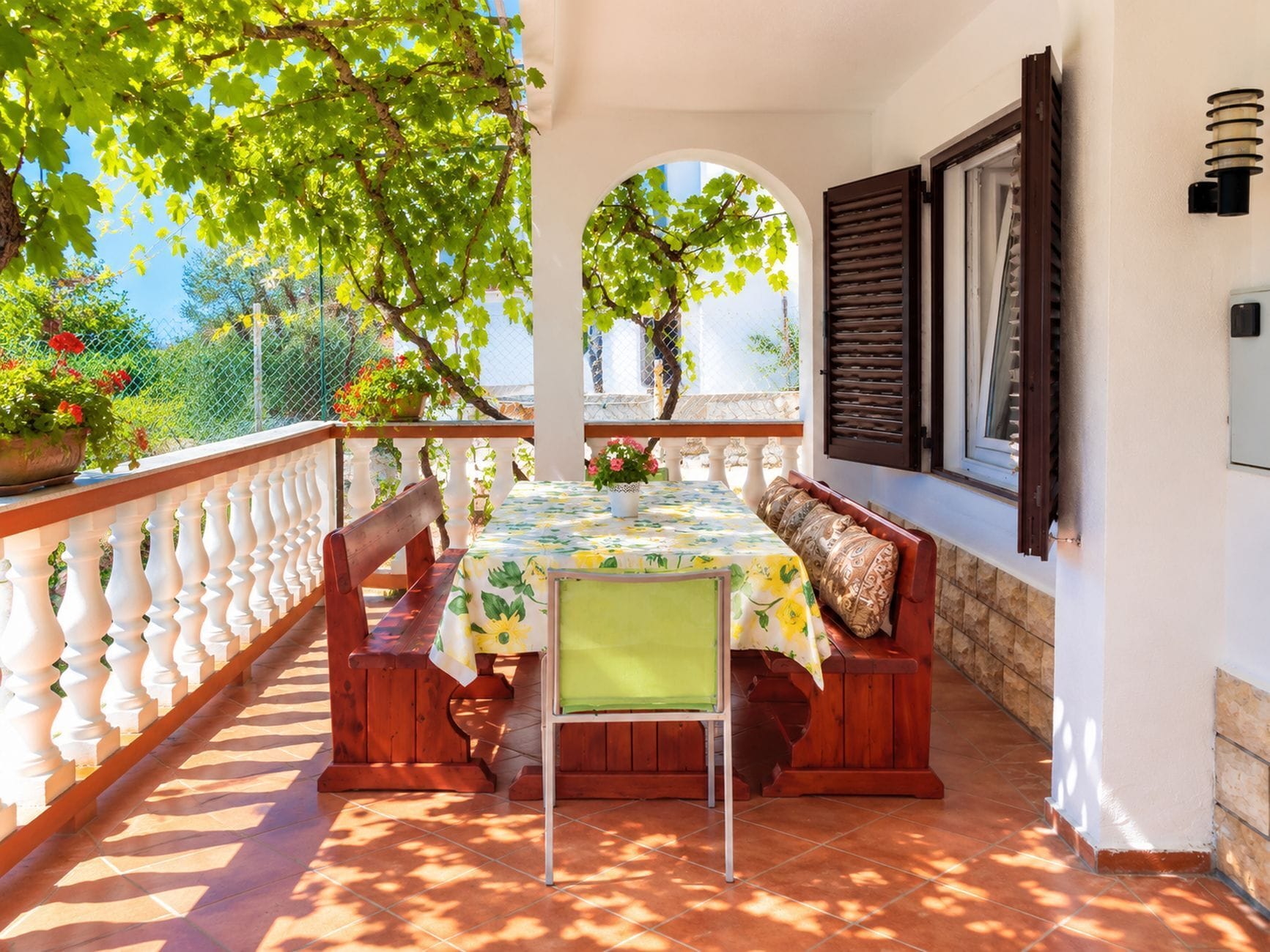 An outdoor patio with a table and benches, surrounded by a white balustrade and a view of greenery.