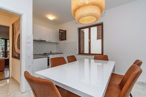 A modern kitchen with white cabinets, a built-in oven, and a dining area with a white table and brown chairs. - Apartments Milan (Lopar)