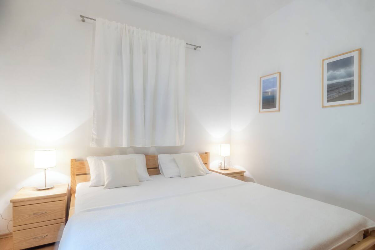 A neatly made bed with white linens, two bedside lamps, a wooden nightstand, and a view of the sea through a window with white curtains.