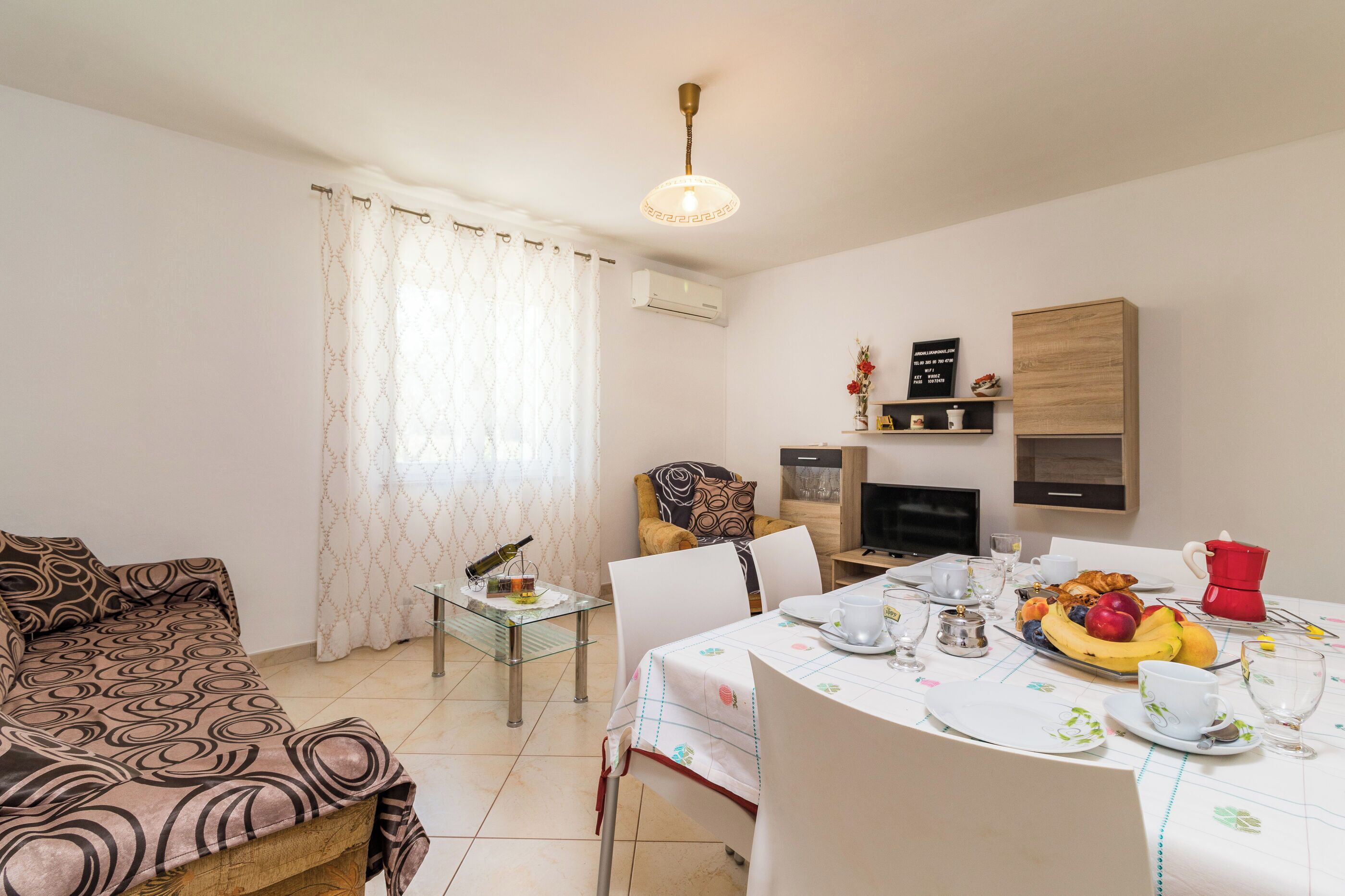 Un salon avec un canapé, une table à manger dressée avec des assiettes, des tasses et un saladier rempli de fruits, et un téléviseur posé sur un meuble.