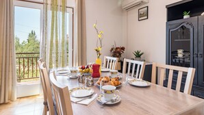 Et spiseområde med et bord dækket til morgenmad, inklusiv tallerkener, kopper og kager. Der er stole omkring bordet, og et vindue med gardiner. En vase med blomster står på bordet, og en plante står på et skab til højre.