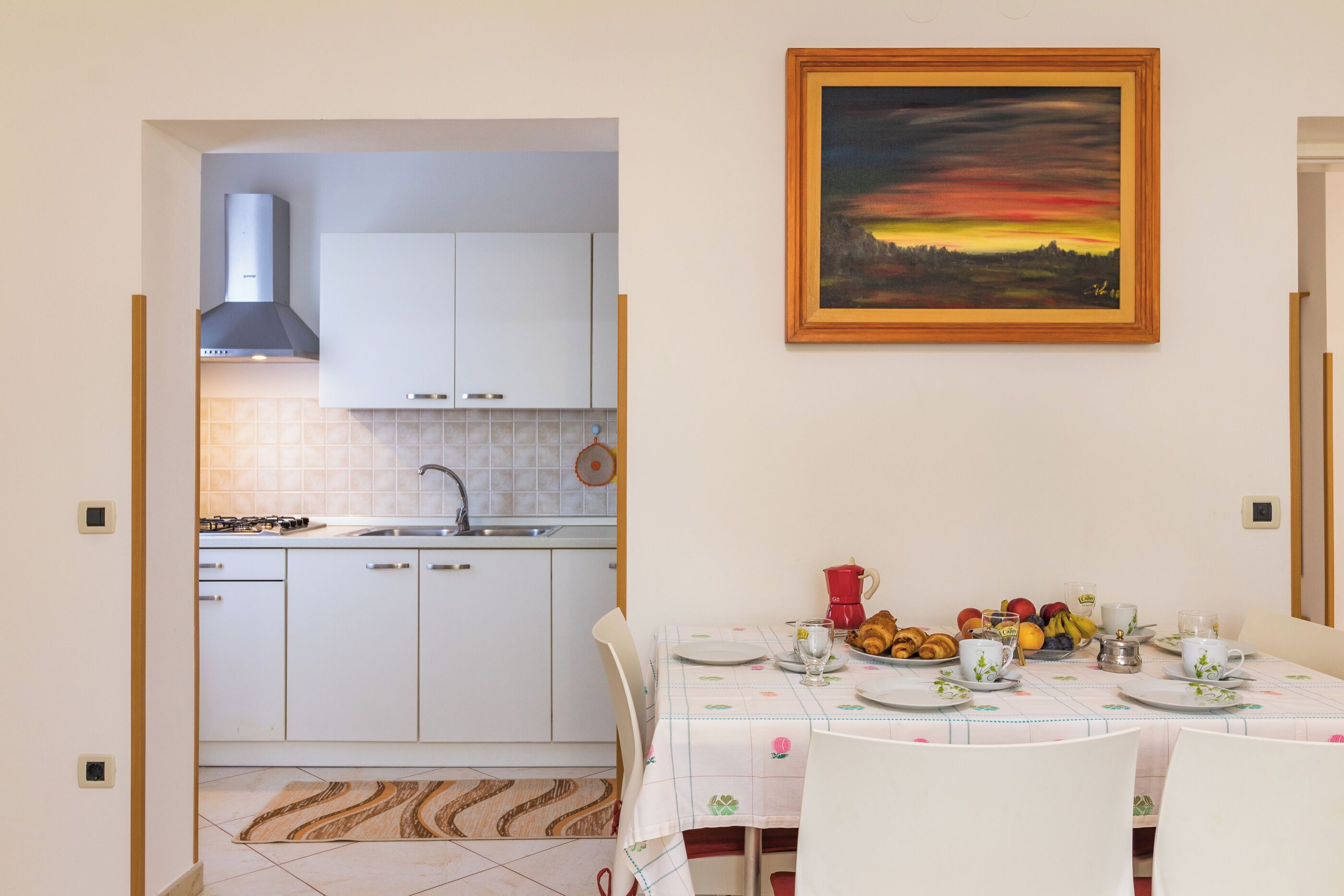 A dining area with a table set for two, a kitchen in the background, and a sunset painting on the wall.