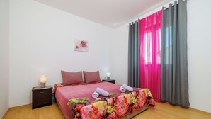 A bedroom with a wooden floor, a bed with a floral bedspread, two pillows, a nightstand with a lamp, and a window with pink curtains. - Apartment Lea (Crikvenica)