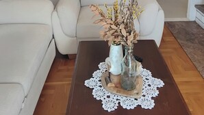 A modern living room with a sofa, a coffee table with a vase of dried flowers, and a kitchen in the background with white cabinets and a refrigerator. - Apartment Anea (Marina)