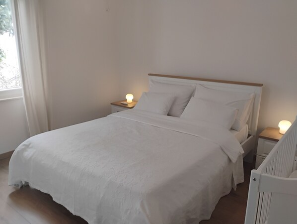 Un lit bien fait dans une pièce aux murs blancs et dotée d’une fenêtre. On y trouve deux tables de chevet avec des lampes.