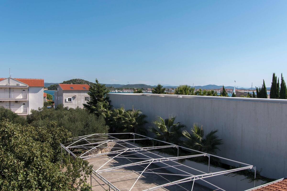 A rooftop terrace with a metal framework, surrounded by greenery and buildings in the background.