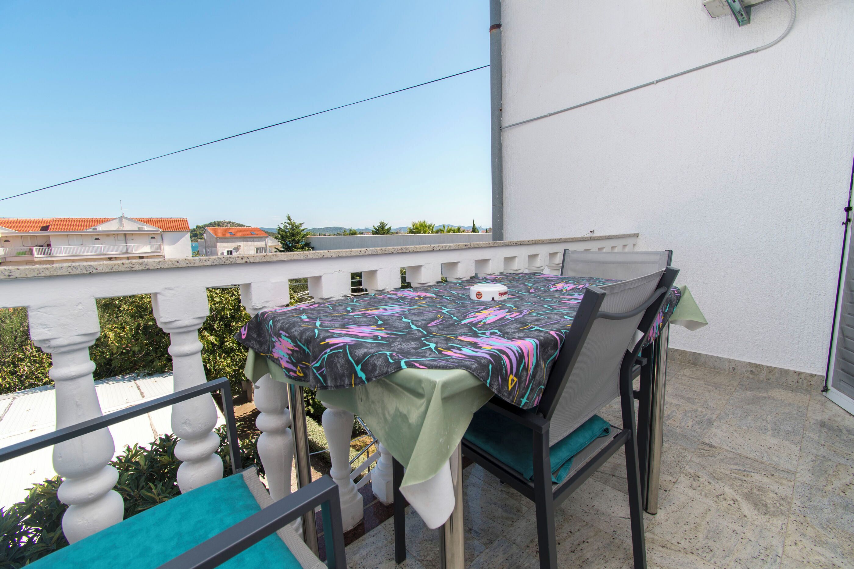 A balcony with a table and chairs, a white railing, and a view of buildings in the background.