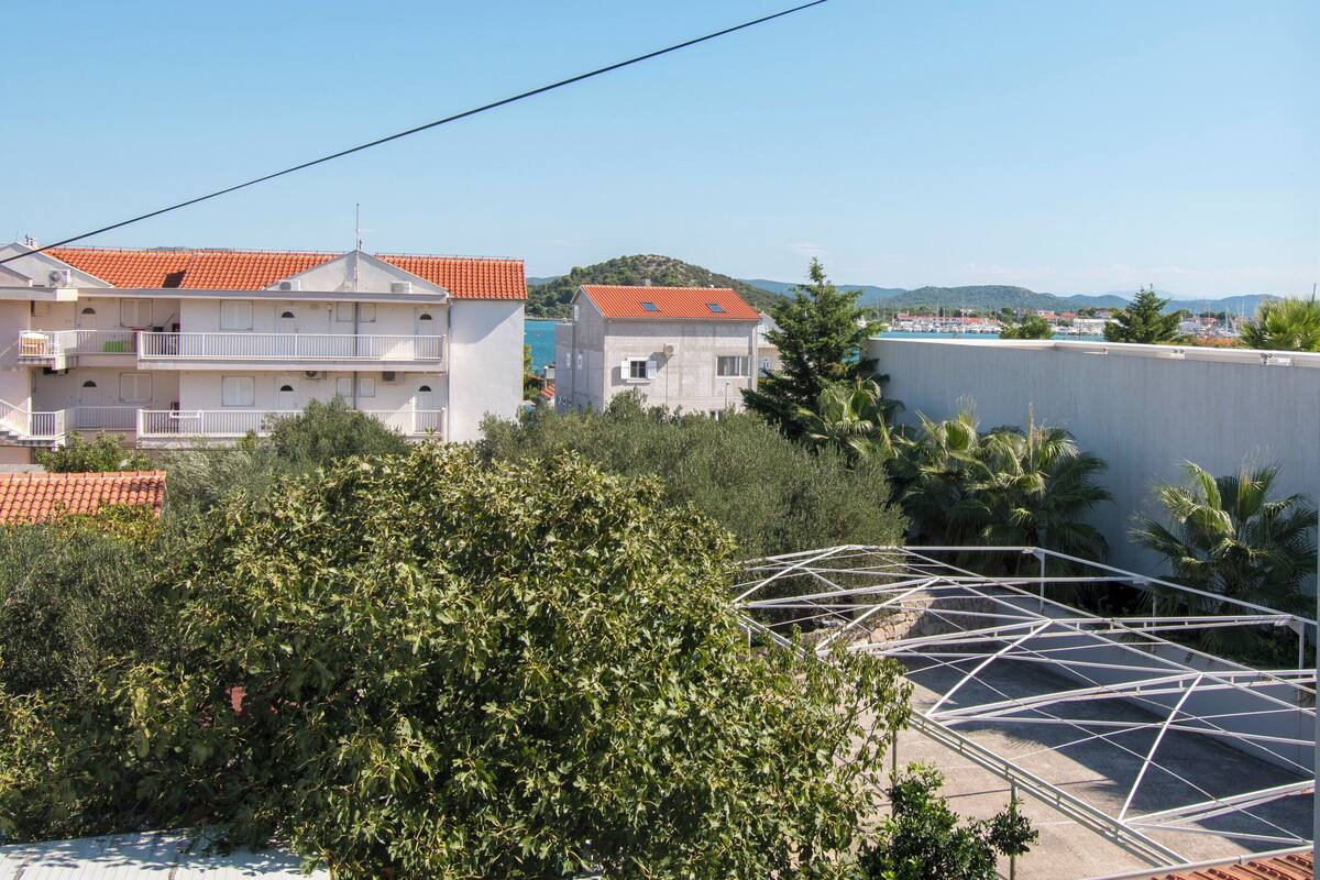 A residential area with buildings featuring red-tiled roofs, a clear blue sky, and a view of the sea.