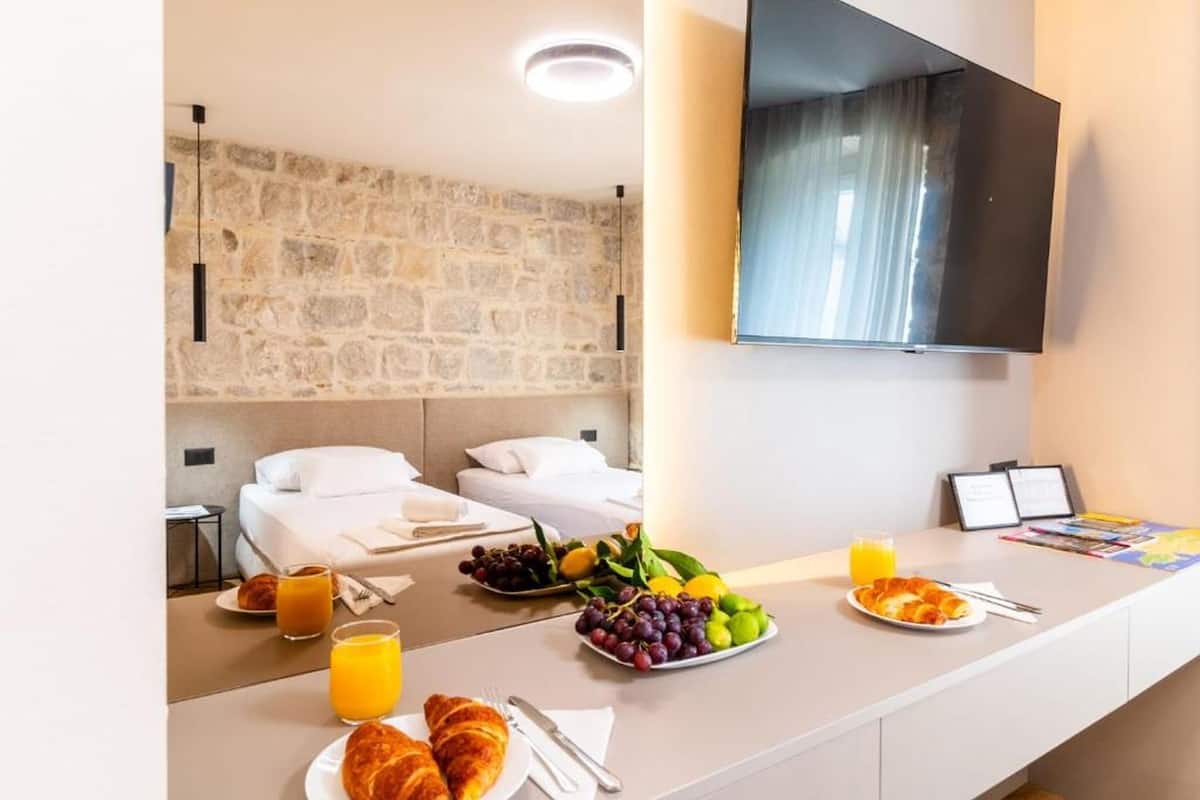 A hotel room with a stone wall, a flat-screen TV, a breakfast table with croissants, fruit, and orange juice.