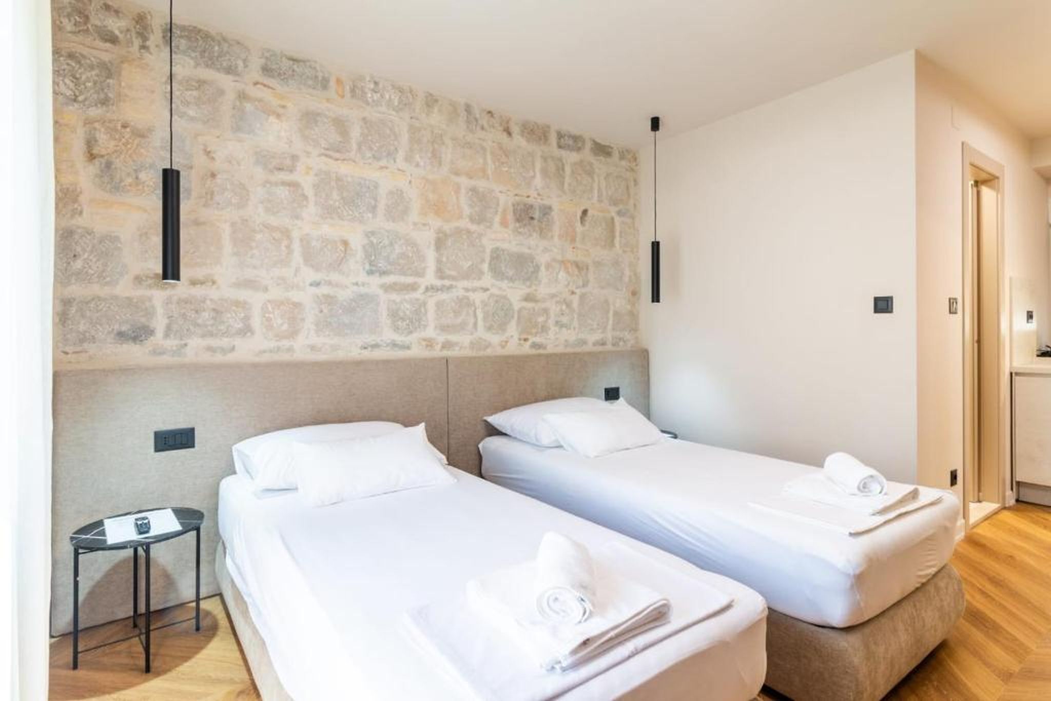 A hotel room with two single beds, a stone wall, a small table, and a door leading to a bathroom.