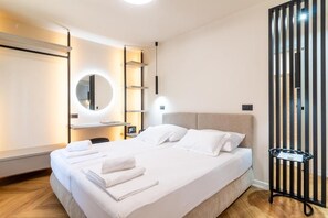 Moderni hotellihuone, jossa on suuri sänky, pyöreä peili, yöpöytä ja musta metallinen hylly.