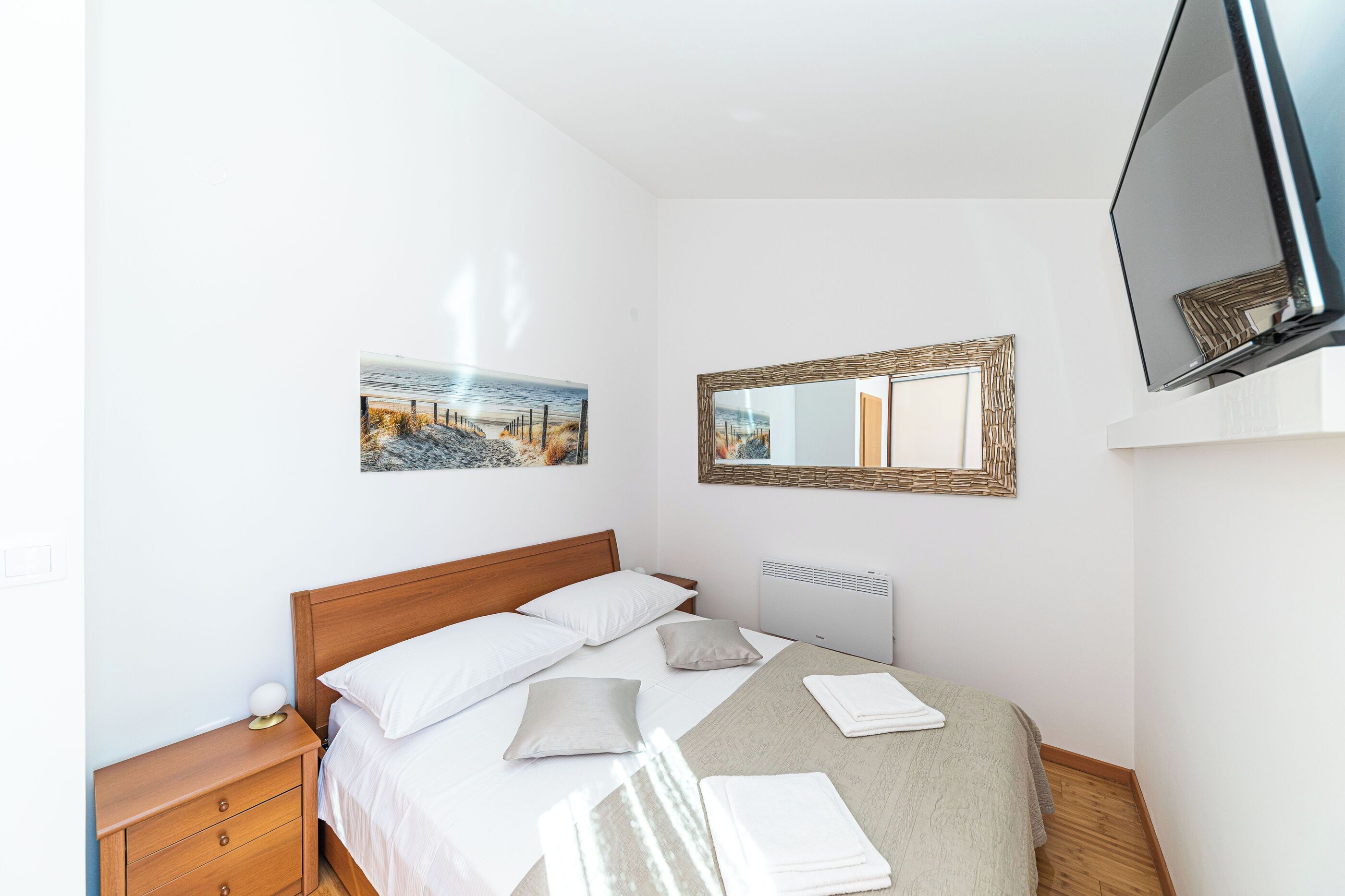 A neatly made bed with pillows and a blanket, a bedside table with a lamp, a wall-mounted TV, and a framed picture on the wall.