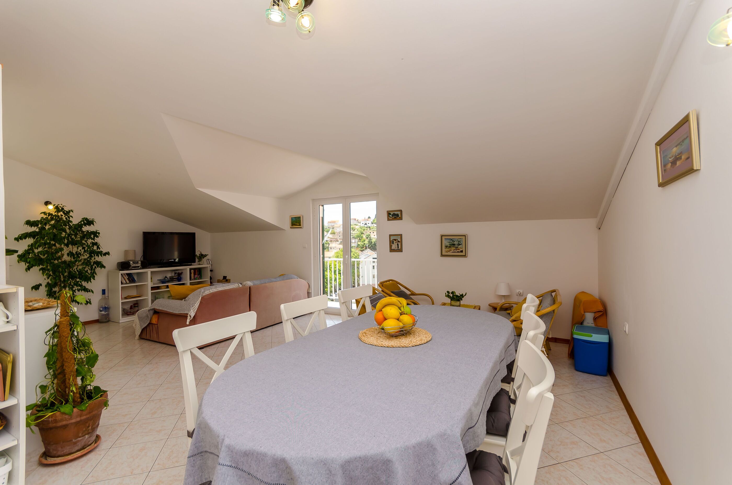 A dining area with a table set for four, a potted plant, and a television in the background.