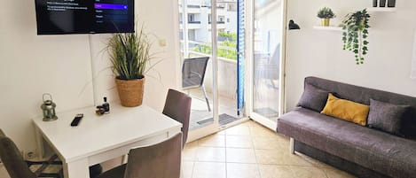 A modern living room with a white table, two chairs, a grey sofa with a yellow cushion, a TV mounted on the wall, and a potted plant.