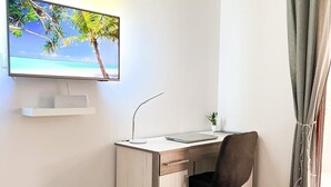 A hotel room with a bed, a desk with a laptop, a chair, a TV displaying a beach scene, and a window with curtains. - Apartments Apartments -T and T (Okrug)