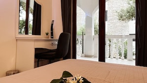 A hotel room with a bed, a desk, a chair, a small plant, and a balcony with a view of a building and greenery. - Apartments Apartments -T and T (Okrug)