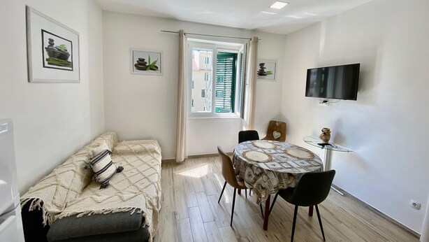 Ett kompakt vardagsrum med en soffa, ett matbord med stolar, en tv som är monterad på väggen och ett fönster med gardiner.