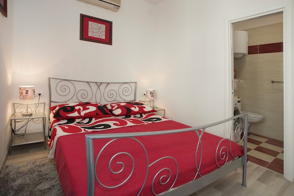 Une chambre à coucher avec un lit en métal, une couvre-lit rouge et noire à motifs, une table de chevet avec une lampe et un tableau encadré au mur.