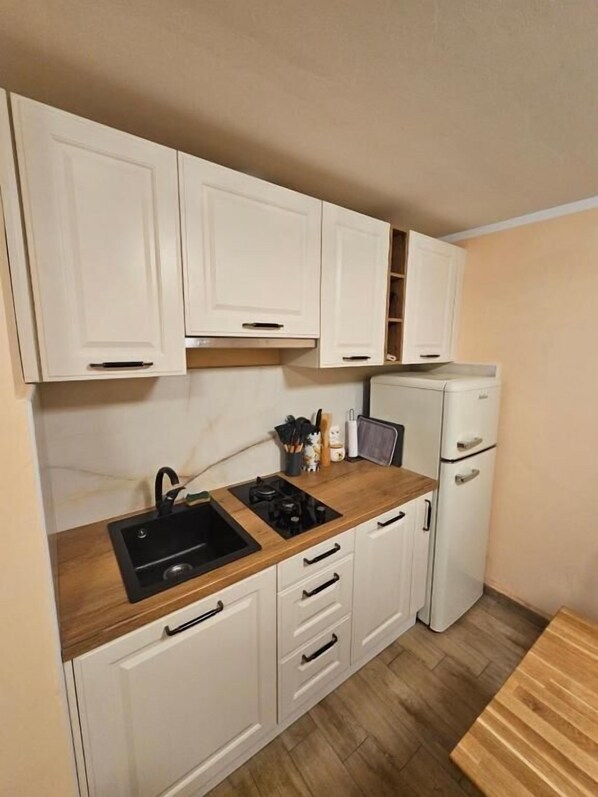 A compact kitchen with white cabinets, a wooden countertop, a black sink, and a white refrigerator. - House Sunshine (Pasman)