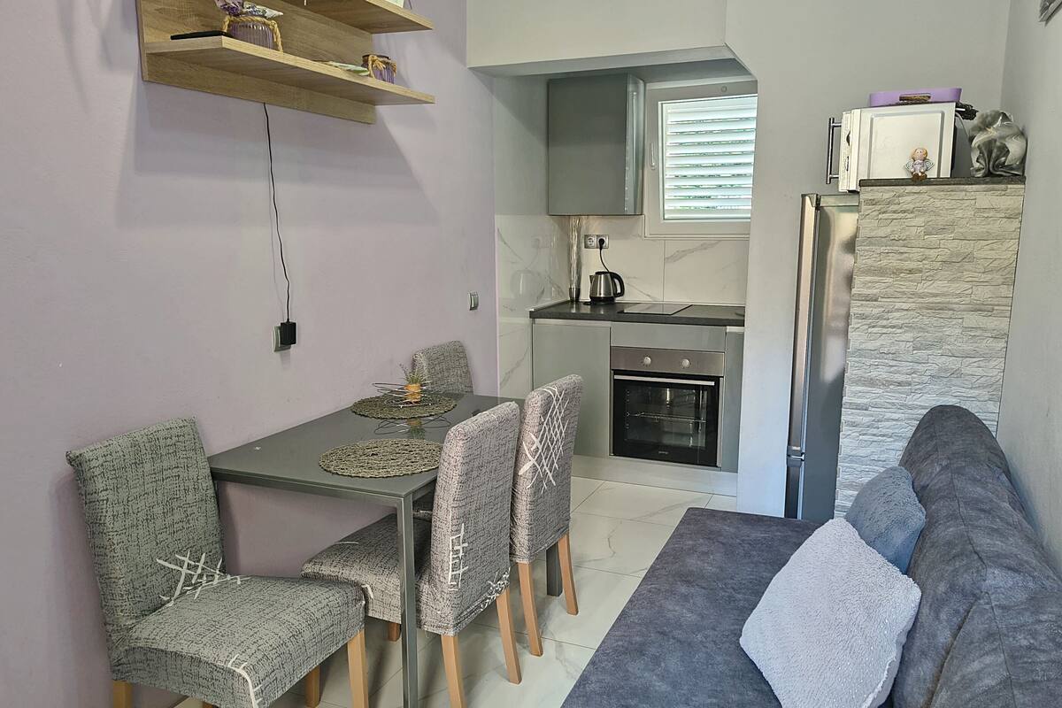 A compact living space with a dining table, four chairs, a sofa, a kitchen area with a refrigerator, oven, and microwave, and a small window with blinds.