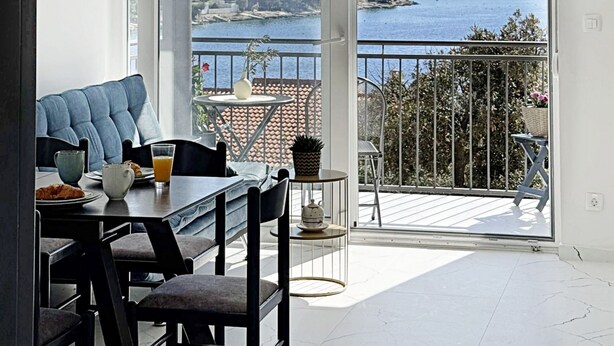 A dining area with a table and chairs, a view of a coastal town, and a balcony with a railing. - Apartment Simun (Rogoznica)