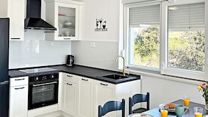 A modern kitchen with white cabinets, a black countertop, and a dining area with a table set for breakfast. - Apartment Simun (Rogoznica)