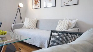 Ein modernes Wohnzimmer mit einem grauen Sofa, einem Glastisch und einem schwarzen Korbsessel. An der Wand befinden sich gerahmte botanische Drucke und eine Stehlampe.