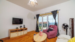 Ein Zimmer mit Holzboden im Fischgrätmuster, einem an der Wand montierten Fernseher, einem Holzkasten mit Schubladen, einem rosa Sessel, einem Bett mit weißer Matratze und einem Standventilator.