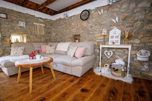 Ein gemütliches Wohnzimmer mit Steinwand, Holzboden, einem beigefarbenen Sofa mit floralen Kissen, einem runden Holztisch mit Spitzenüberzug und einer kleinen weißen Häuserdekoration auf einem weißen Sockel.
