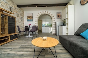 A modern living room with a stone wall, a wooden coffee table, a grey sofa, and a dining area with chairs and a table. - Apartments Villa Magnolia (Mali Losinj)