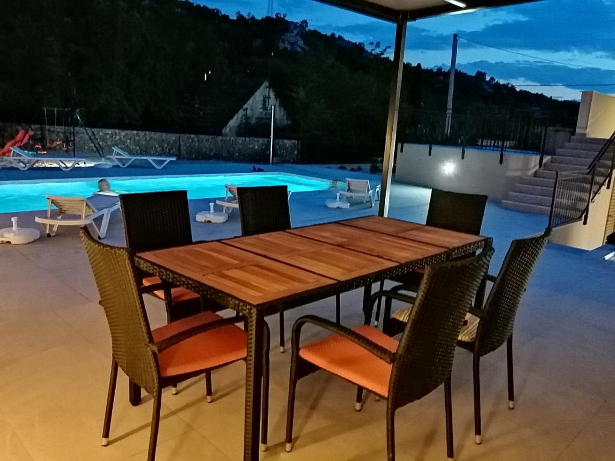 Un espace piscine extérieur avec une table et des chaises en bois, entouré de fauteuils de relaxation et d’un mur en pierre.