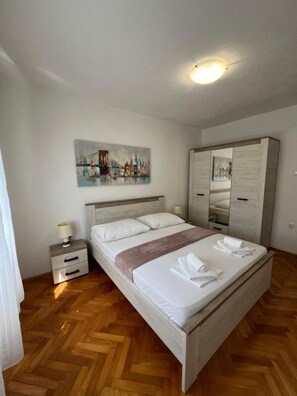 Hotellihuone, jossa on puulattia, valkoisilla lakanoiden peitetty sänky, yöpöytä lampun kanssa, seinällä oleva taulu ja vaatekaappi.