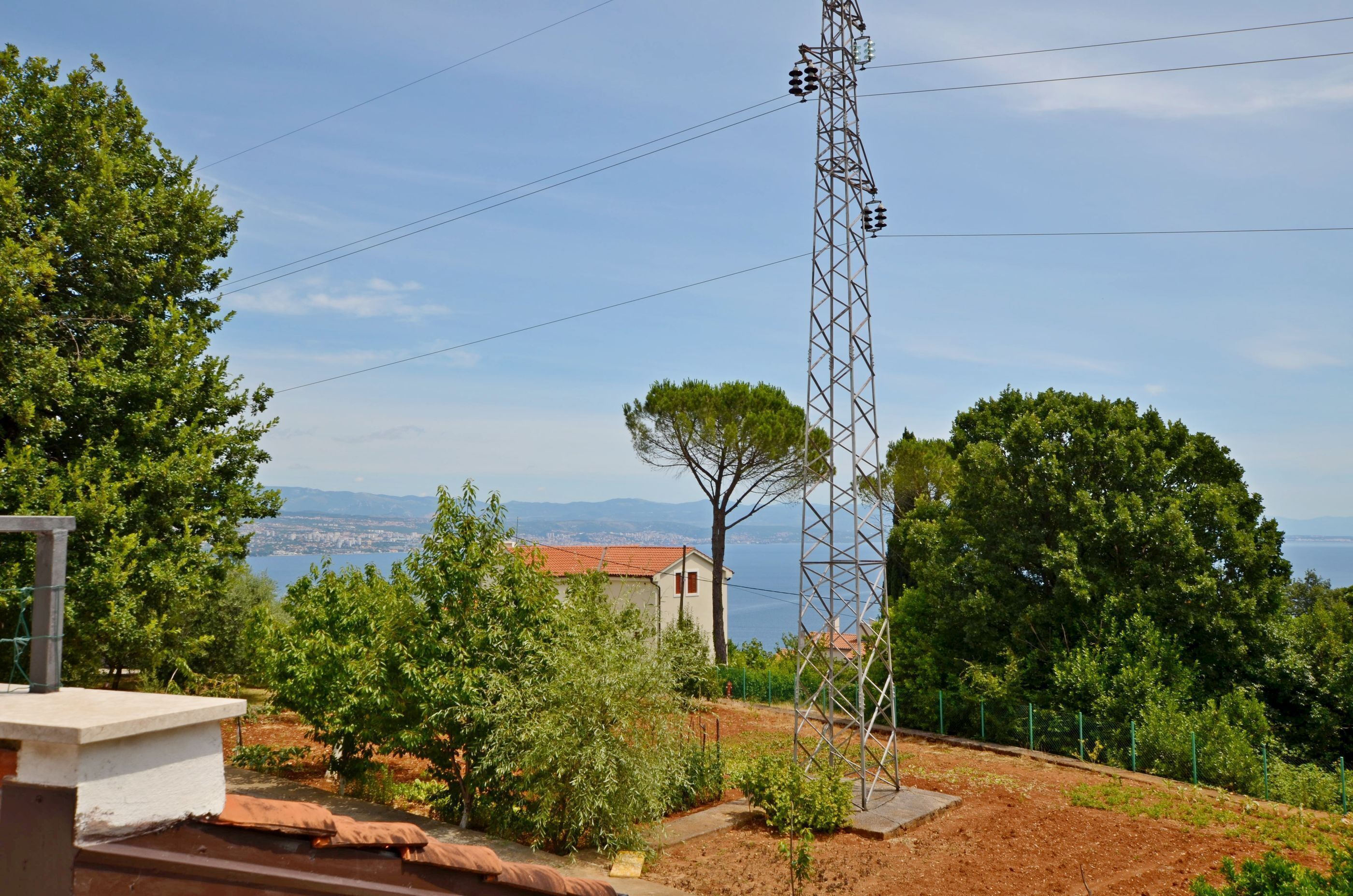 A view of a coastal landscape with a tall communication tower, trees, and a distant view of the sea and mountains.
