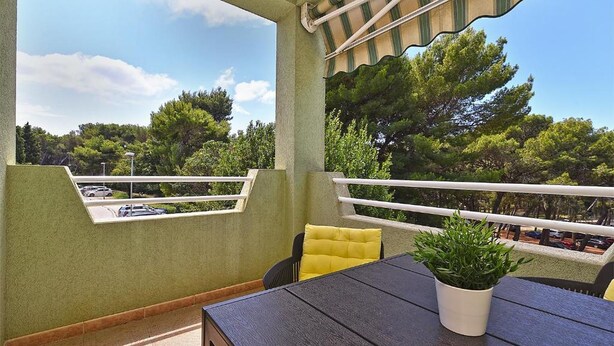 A balcony with a table and chairs, a potted plant, and a view of trees and parked cars. - Apartment Karolina (Pula)