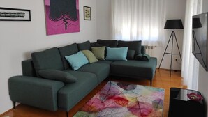 A modern living room with a green sectional sofa, colorful abstract rug, and a large window with white curtains. - Apartment Karolina (Pula)