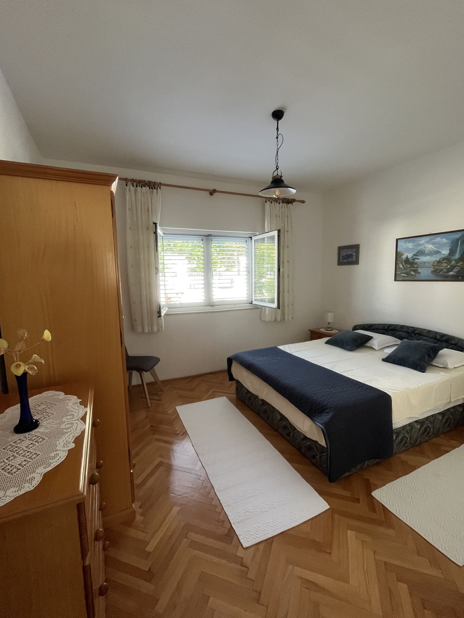 A bedroom with a bed, a wooden wardrobe, a window with curtains, a painting on the wall, and a wooden floor.