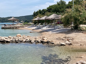 A beach with clear water, rocky shore, and several thatched-roof umbrellas. - Apartment Seasalt (Tisno)