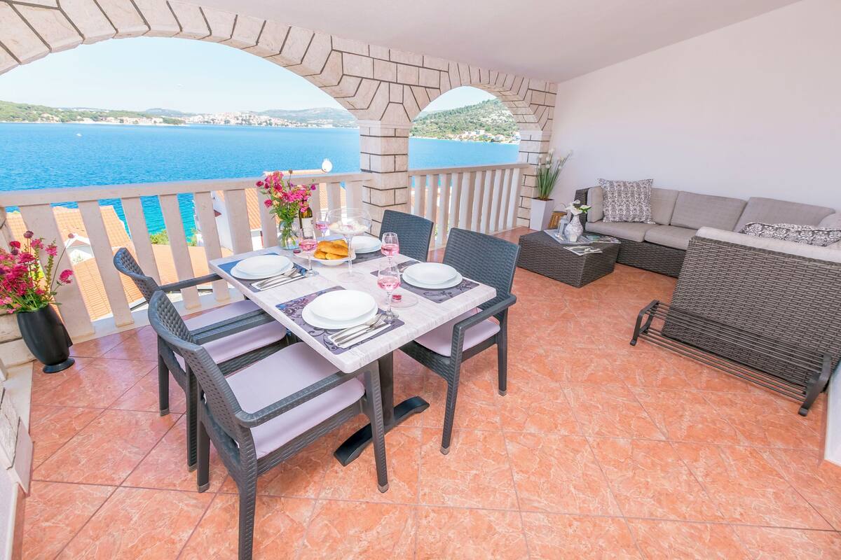 A balcony with a table set for two, chairs, a sofa, and a view of the sea.