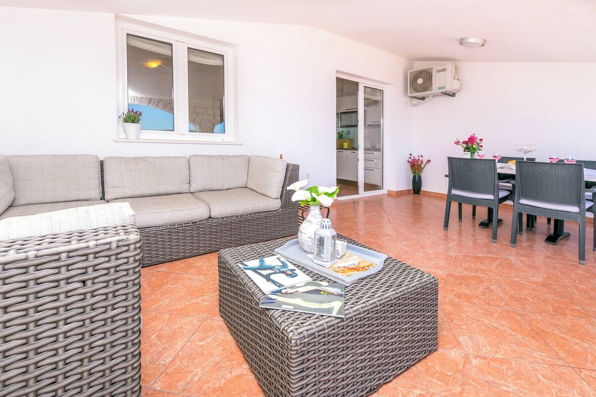 A living area with a sofa, a wicker ottoman, a dining table with chairs, and a kitchen in the background.
