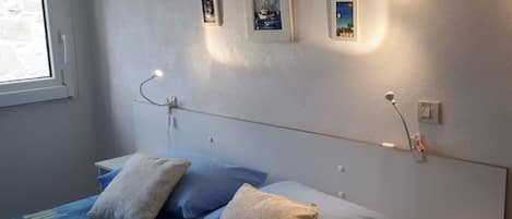 A bedroom with a bed, pillows, a bedside table, a nightstand, a window, and wall-mounted lights.