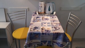 Een eethoek met een tafel bedekt met een tafelkleed met nautische motieven, twee metalen stoelen, een wit schap erboven met een pak tissues, en een koffiezetapparaat.