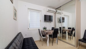 A compact living space with a sofa, a dining table with chairs, a TV mounted on the wall, and a sliding glass door leading to another room. - Apartment Roli (Crikvenica)