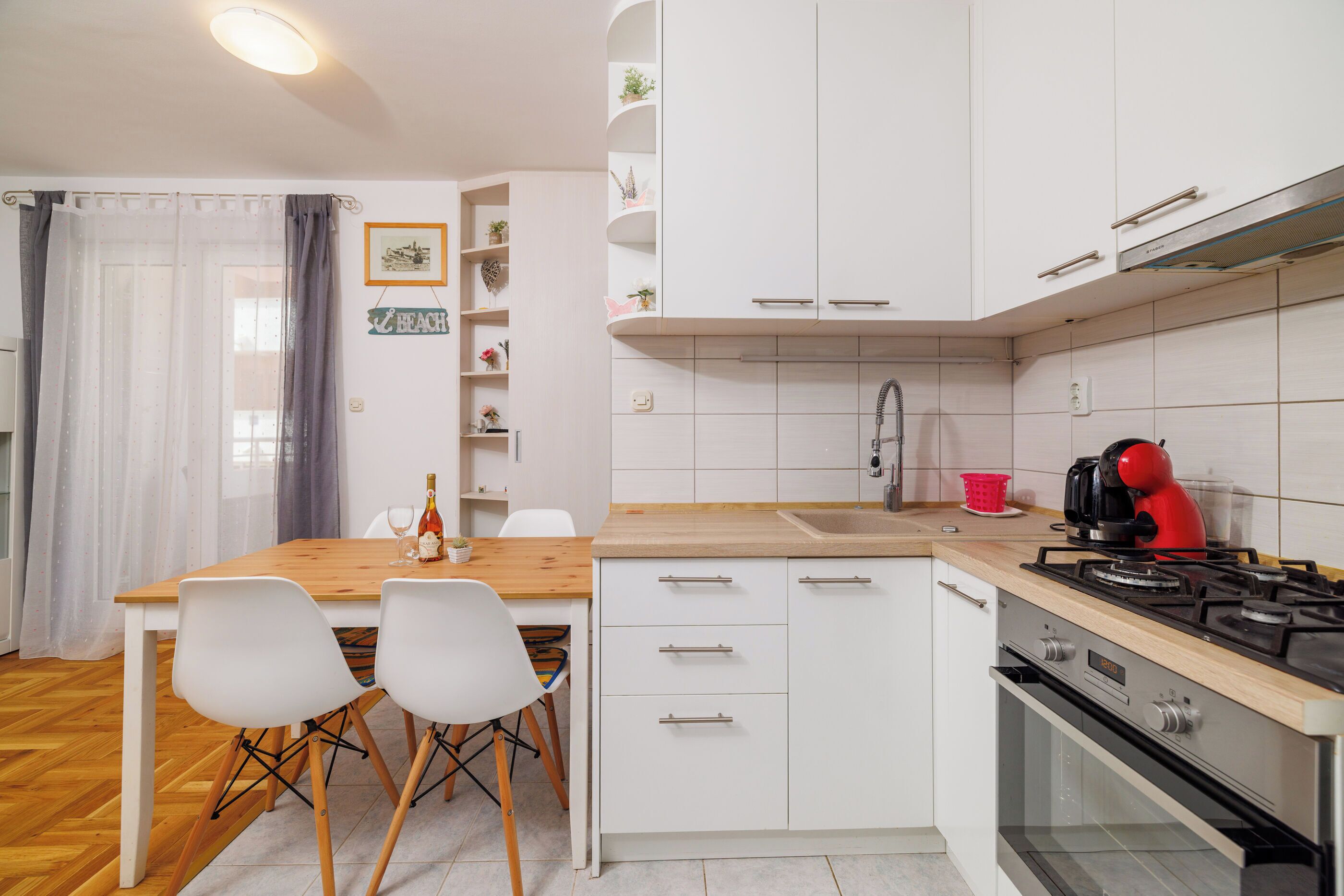 A compact kitchen with white cabinets, a wooden countertop, a gas stove, and a sink. There are two white chairs at a small table, and a bottle of wine is visible on the countertop.