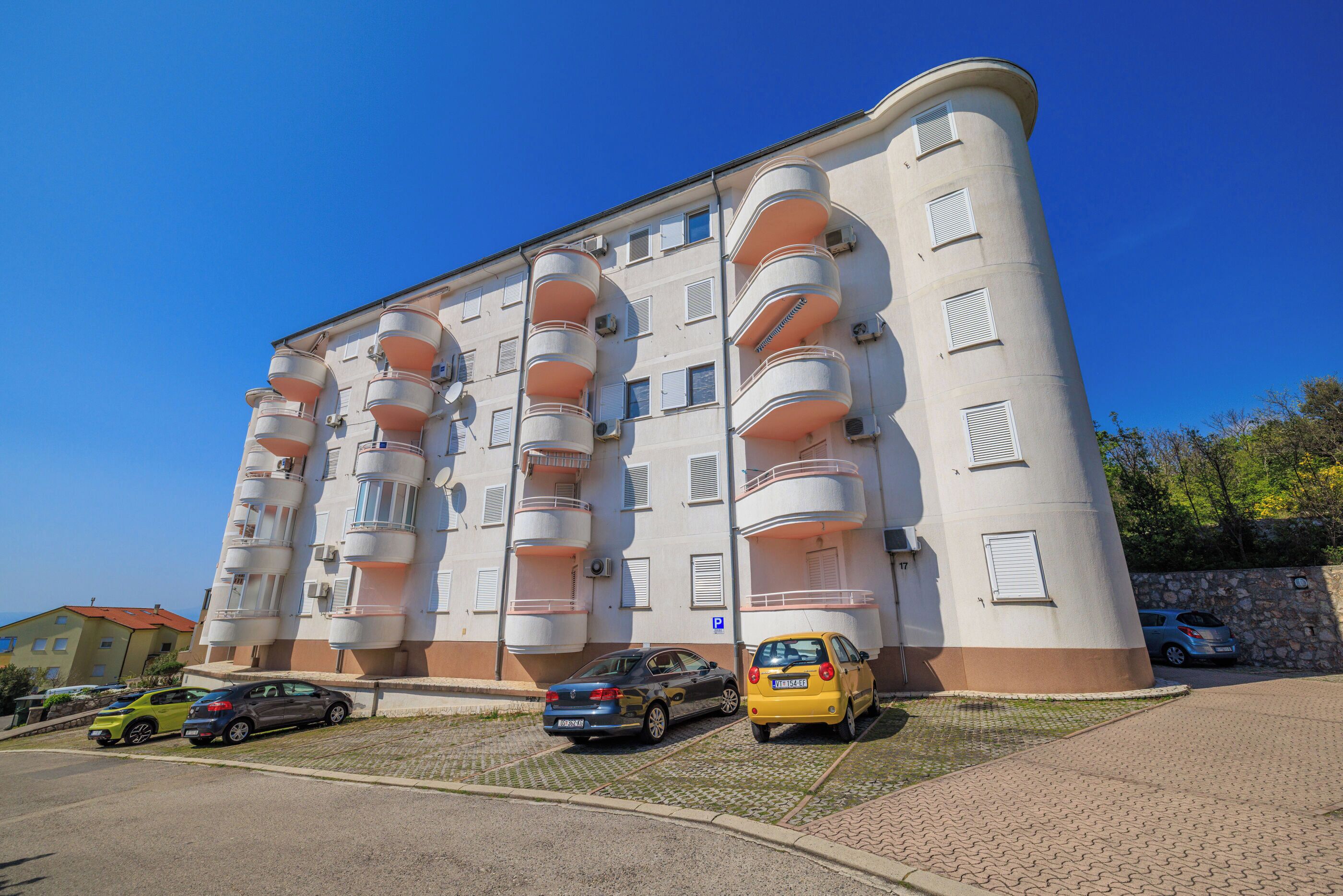 A multi-story residential building with balconies, a parking area, and a clear blue sky.
