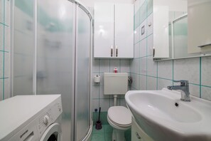 A bathroom with a washing machine, toilet, sink, and glass shower enclosure. - Apartment Roli (Crikvenica)