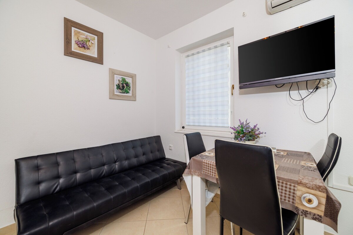 Ein kleiner Wohnbereich mit einer schwarzen Couch, einem Esstisch mit Stühlen, einem flachbildfernseher an der Wand und zwei gerahmten Bildern.