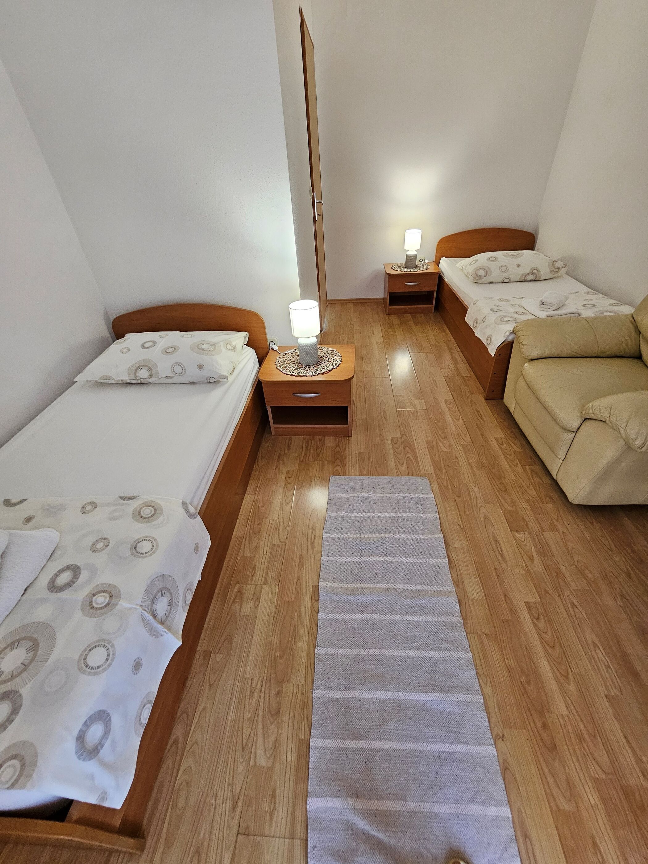 A hotel room with two single beds, a wooden floor, a small bedside table with a lamp, a sofa, and a large mirror.