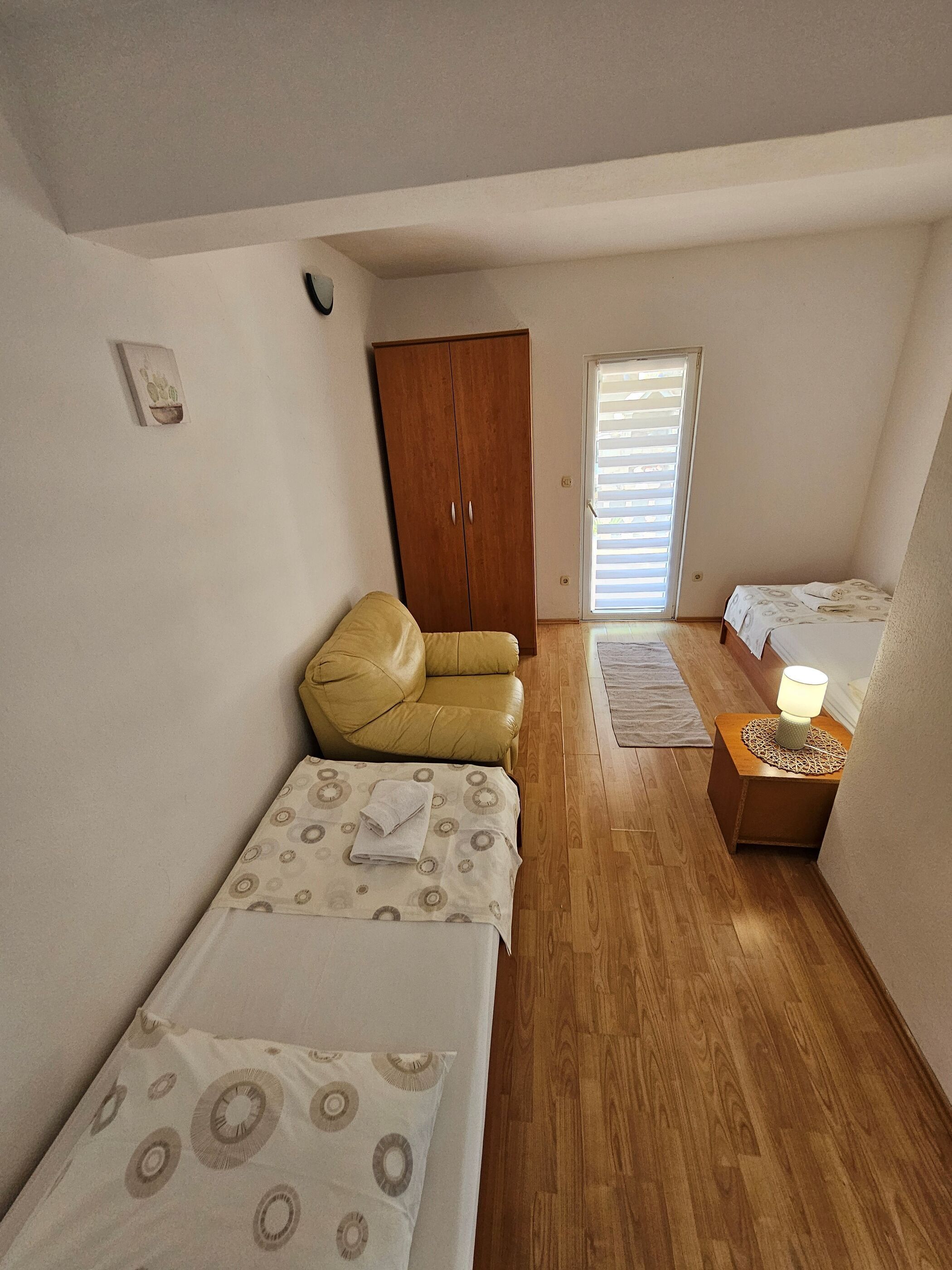 A small, well-lit bedroom with a single bed, a yellow chair, a wooden wardrobe, and a bedside table with a lamp.